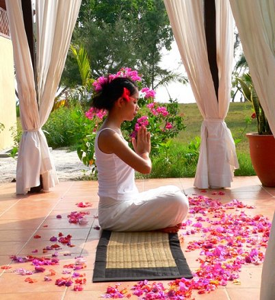 Woman in white seated on a mat with her hands pressed together, flower petals scattered on the floor of an open-air pavilion, Jamu Spa, Mataking Island